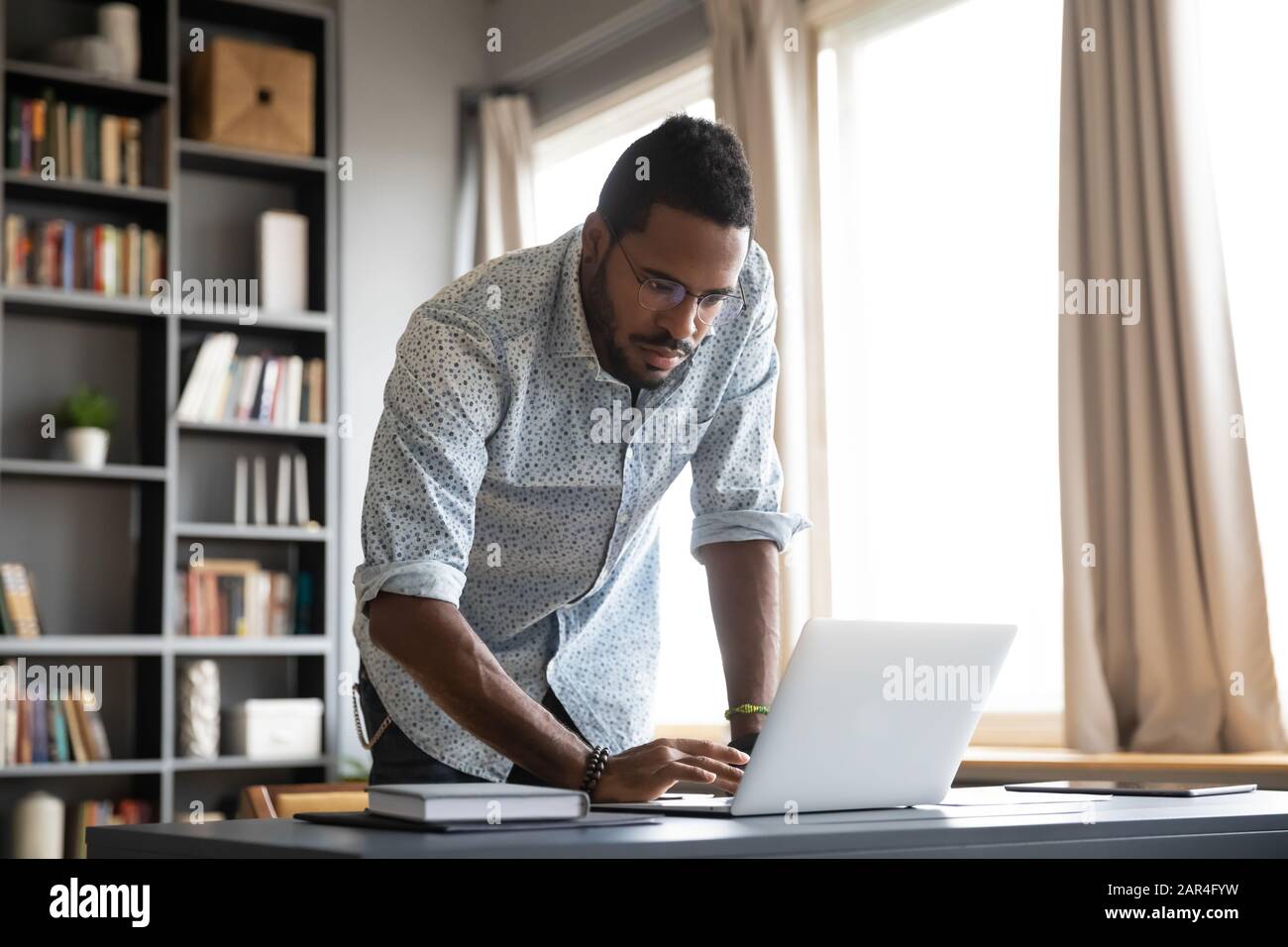 Focused african businessman standing at home office desk using laptop Stock Photo