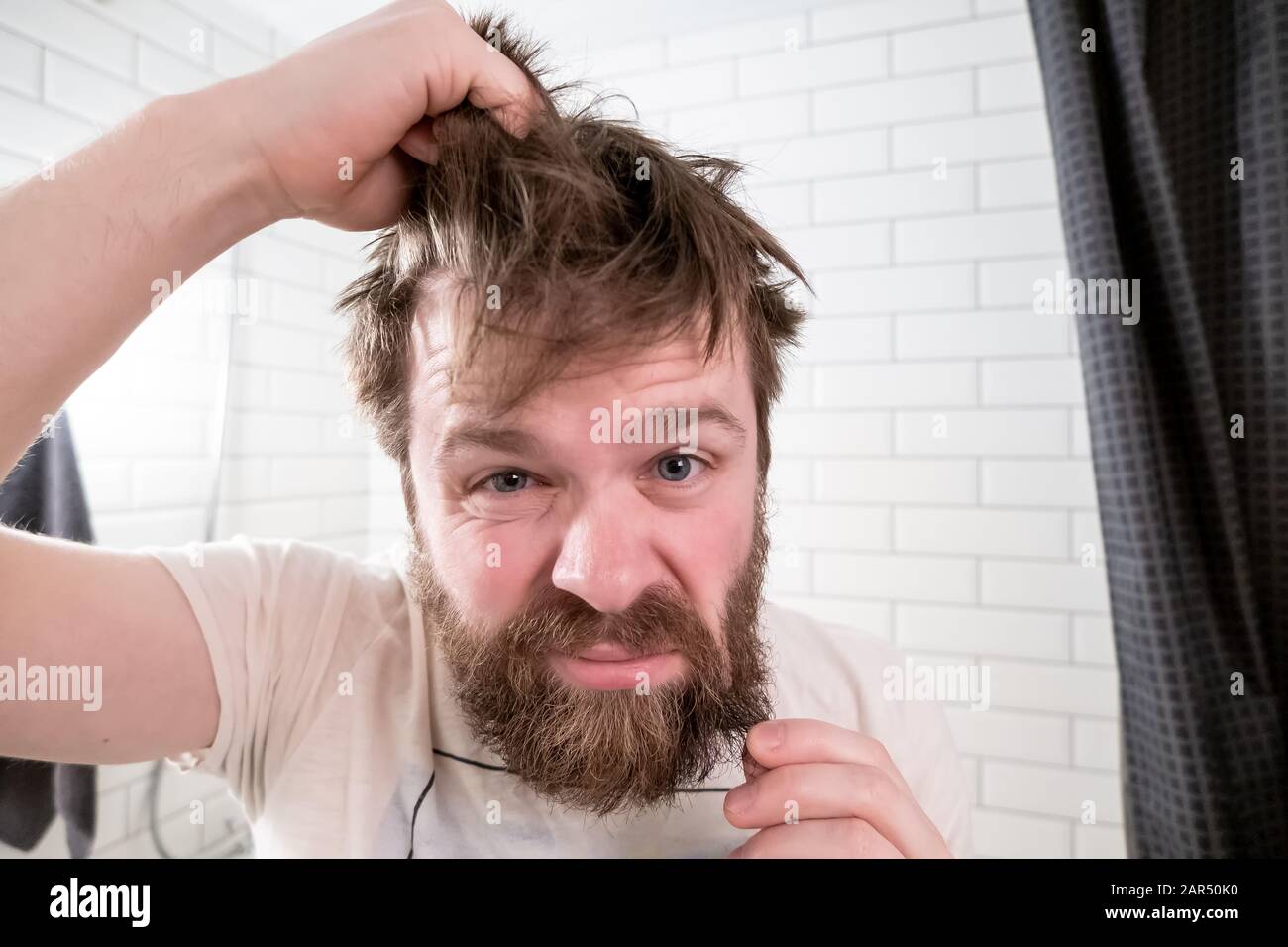 Overgrown man, with a shaggy hairstyle and beard, looks in his reflection in the mirror and is dissatisfied with his appearance. Stock Photo