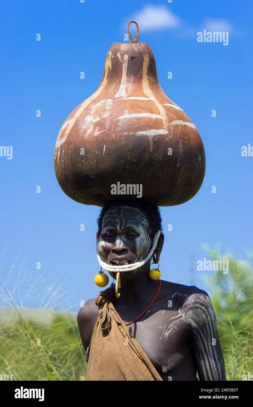 The Mursi women carrying a  big jar on his head. The reason of this 'ornament' is for avoiding to be caught as slaves. Mursi village, Ethiopia Stock Photo