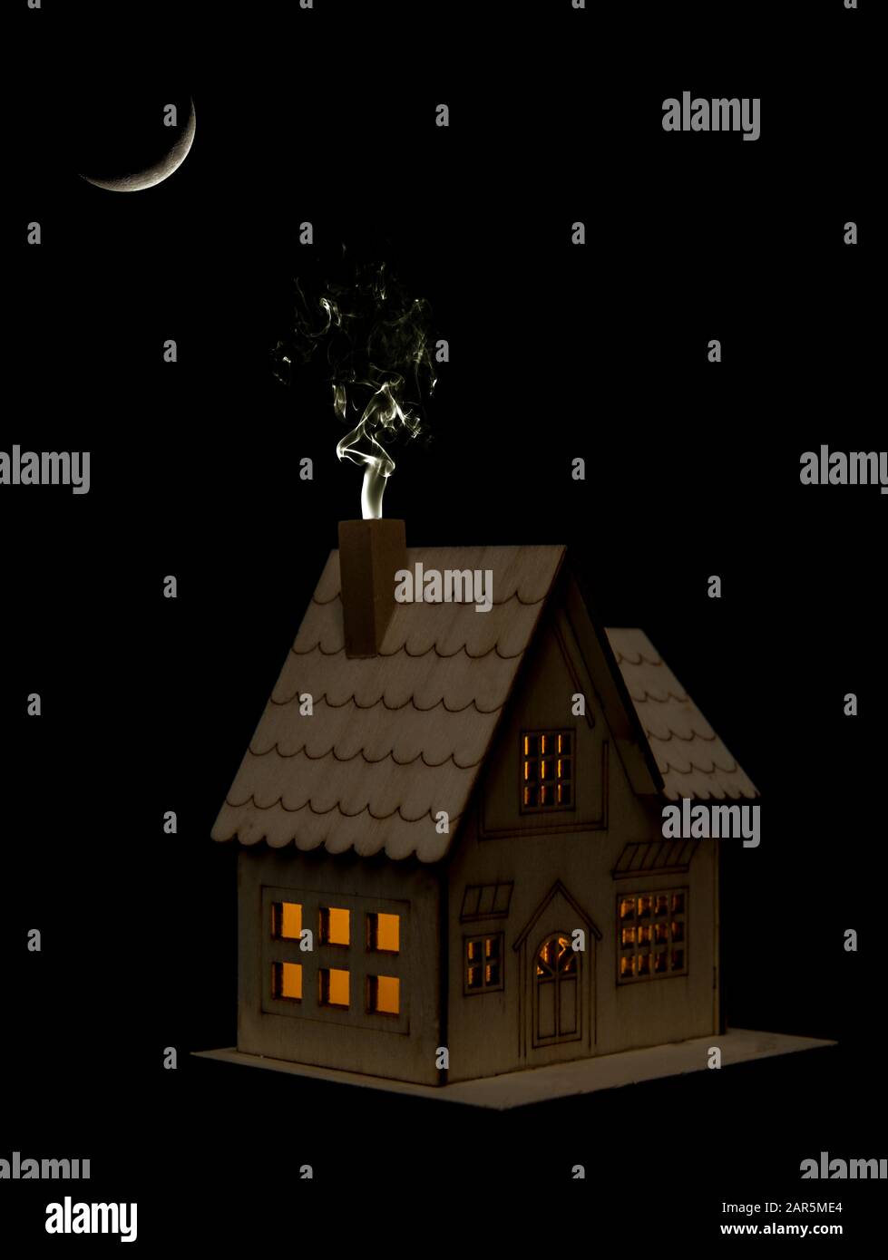 Toy house with lights on at night under a crescent moon, smoke from chimney. Leaving the lights on, waste of electricity, power, not eco friendly. Stock Photo