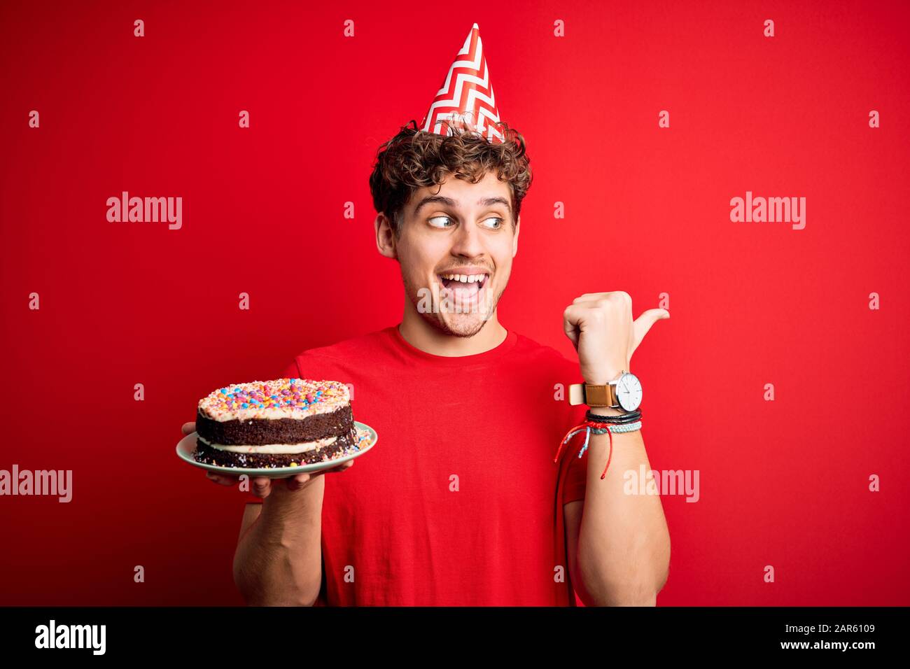 Young blond man with curly hair wearing birthday hat holding cake over ...