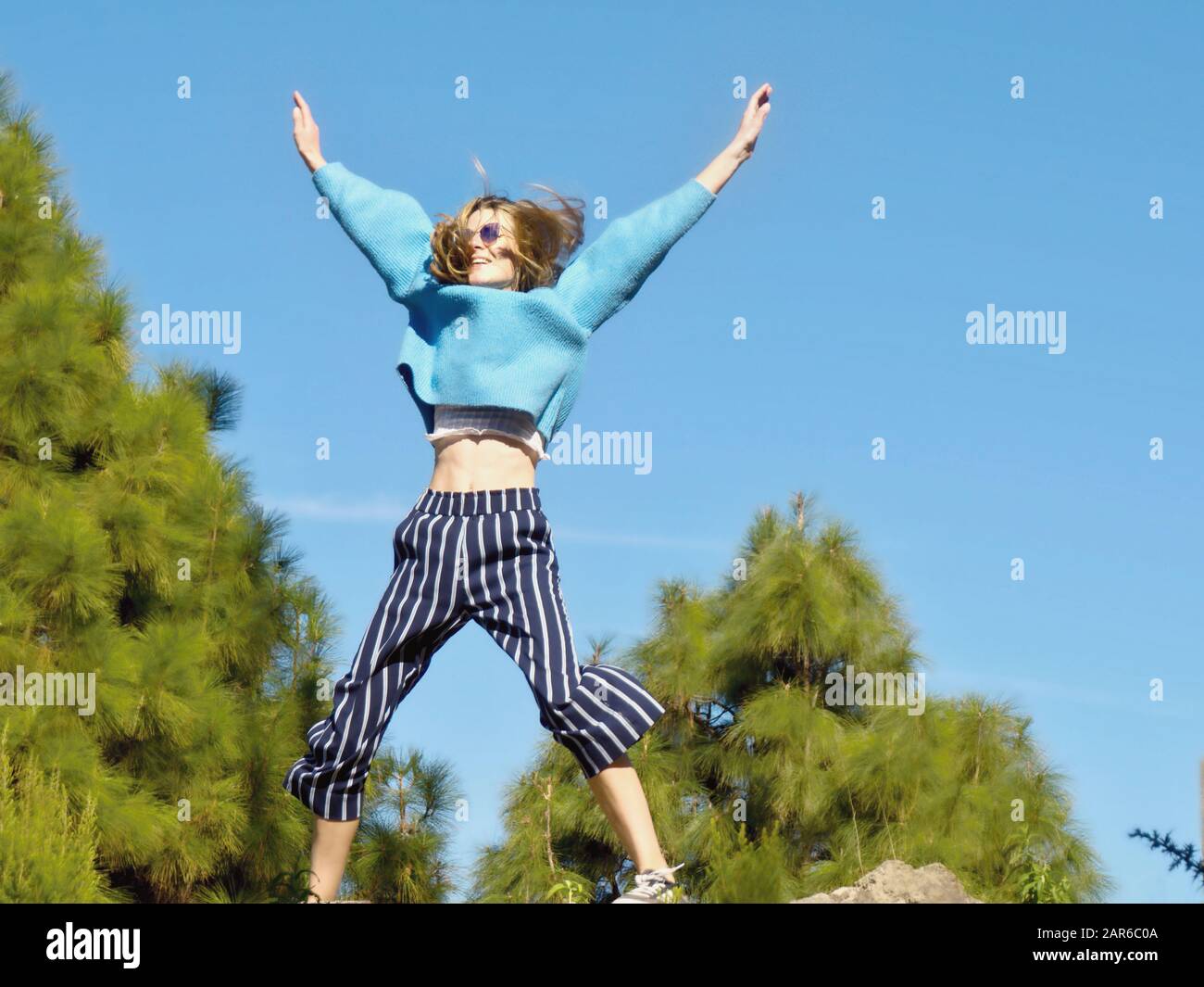 A young woman with a blue sweater and dark blue striped pants makes a leap in the air, her body forming an X shape. Behind it a few green trees and ve Stock Photo