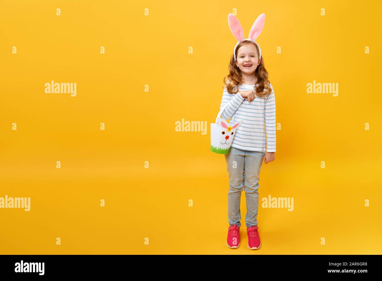 A little girl is standing and holding a bag for Easter eggs and sweets. Happy child in full-growth in bunny ears on a yellow background. Stock Photo