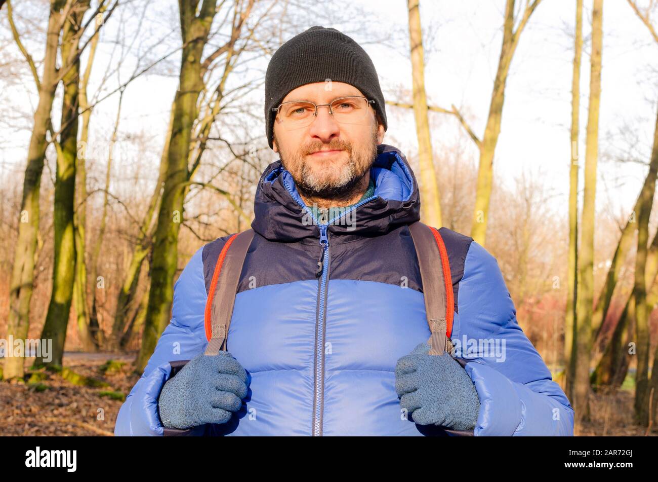 A portrait of a middle-aged man in blue down jacket and a backpack in the barren forest on a sunny day Stock Photo