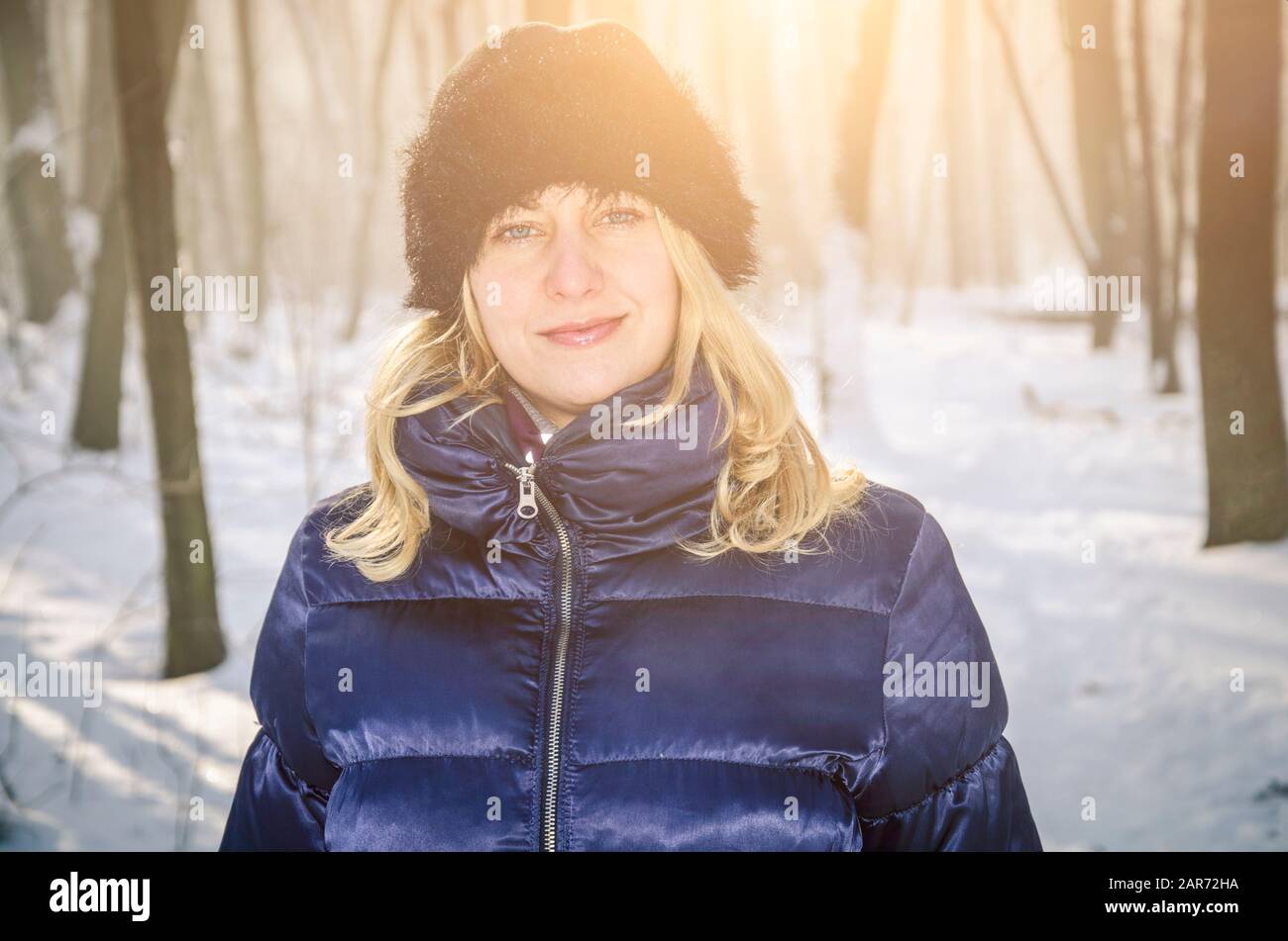 A portrait of a blonde woman in a blue down jacket and fur cap in the snowy winter forest Stock Photo