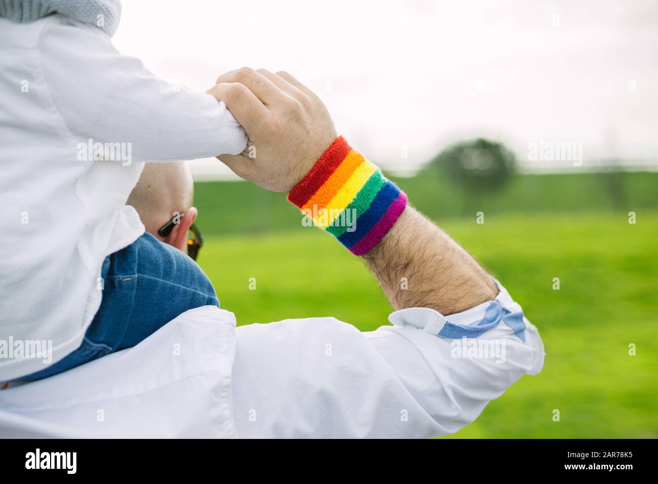 Adult man wearting a gay pride rainbow flag wristband and carrying his adopted son in the park. LGTB and equality concept Stock Photo