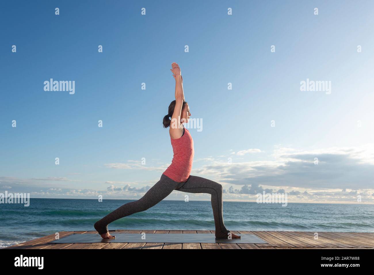 Sporty woman in sportswear practicing yoga, standing in anjaneyasana pose, idyllic location by the ocean Stock Photo