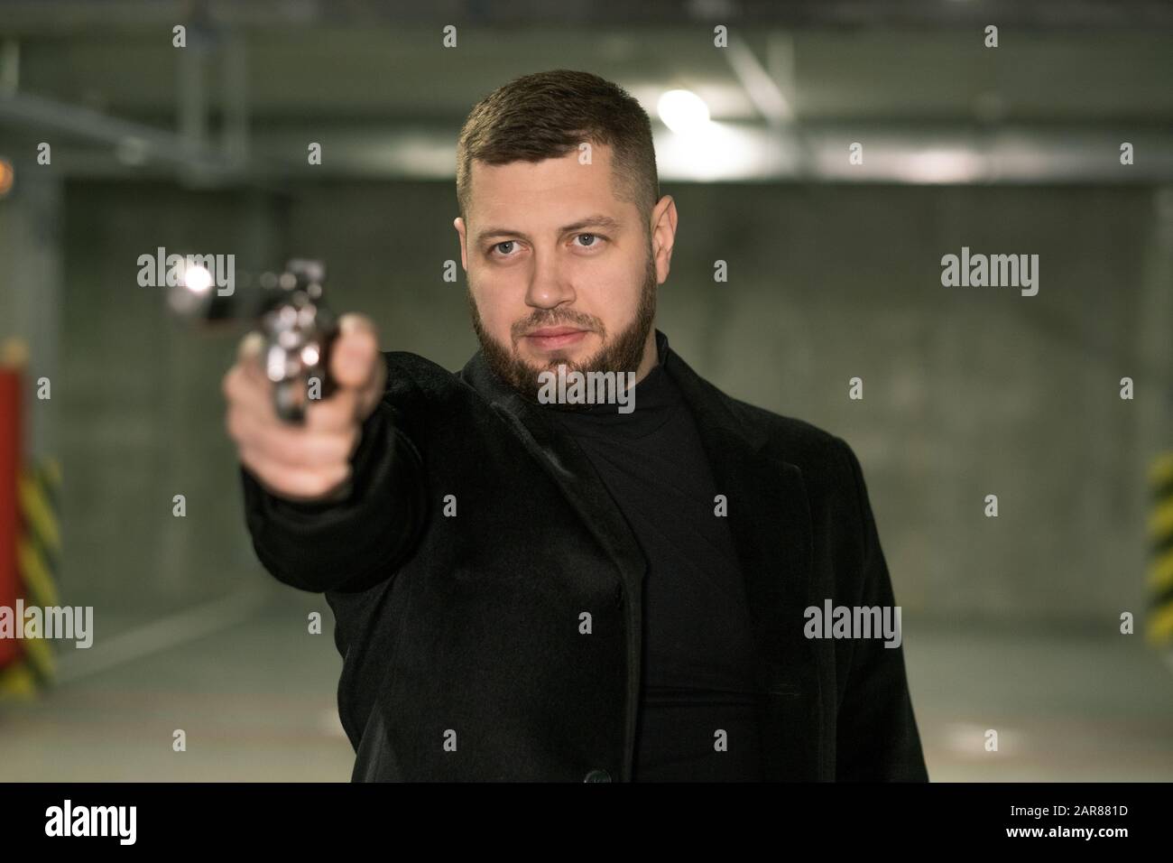 Young killer or agent in black jacket and t-shirt directing handgun on victim Stock Photo