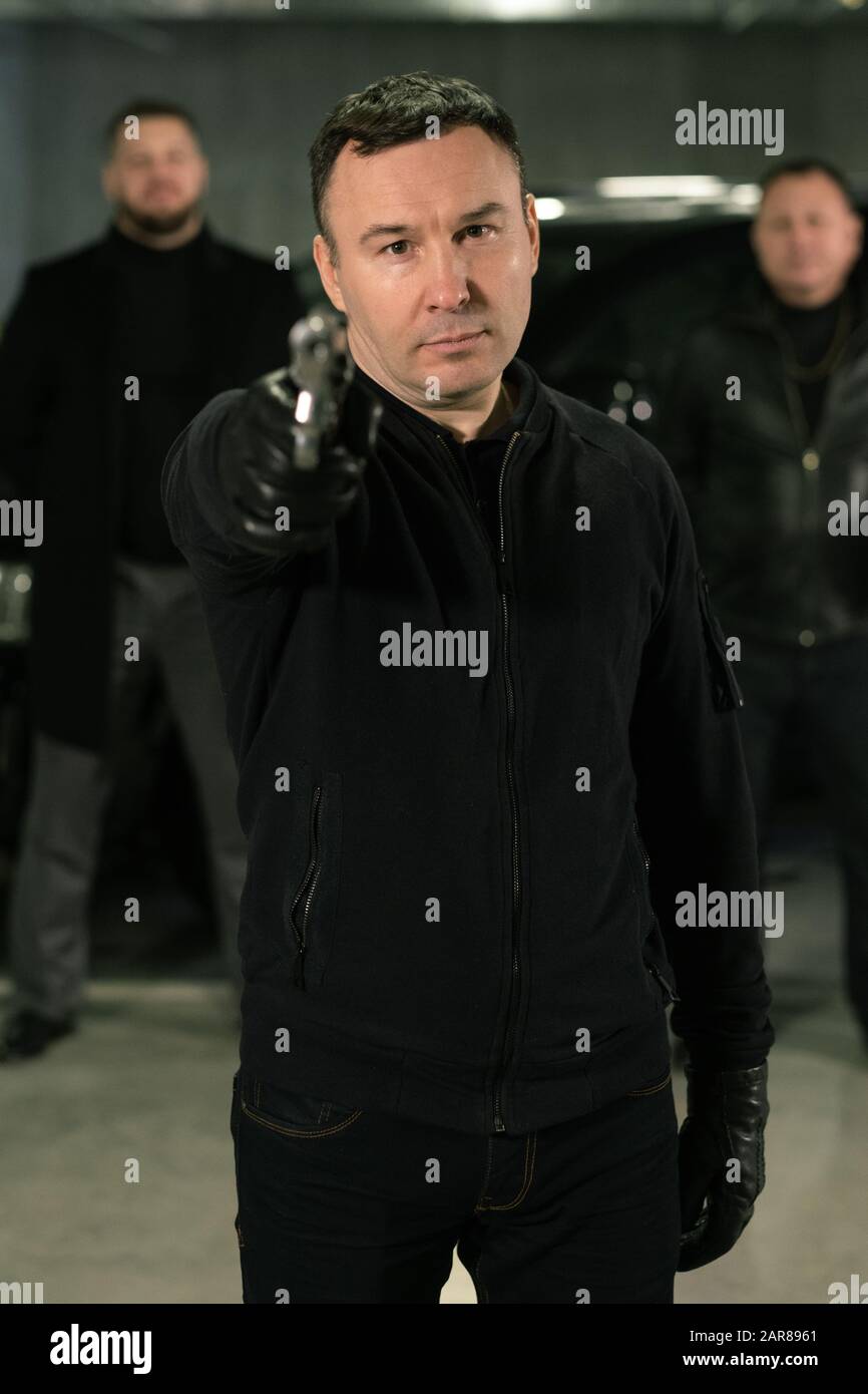 Terrorist in black jeans, jacket and leather gloves pointing at you with handgun Stock Photo