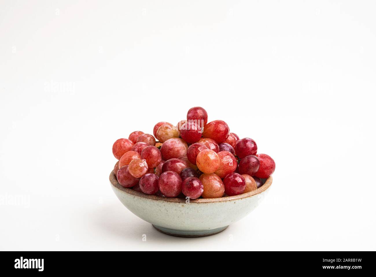 A bunch of fresh red grapes bunched on a small ceramic bowl set on a plain white background. Stock Photo