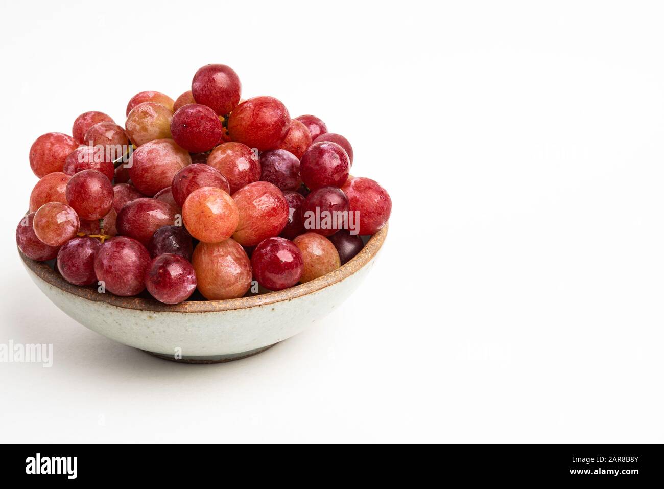 A bunch of fresh red grapes bunched on a small ceramic bowl set on a plain white background. Stock Photo