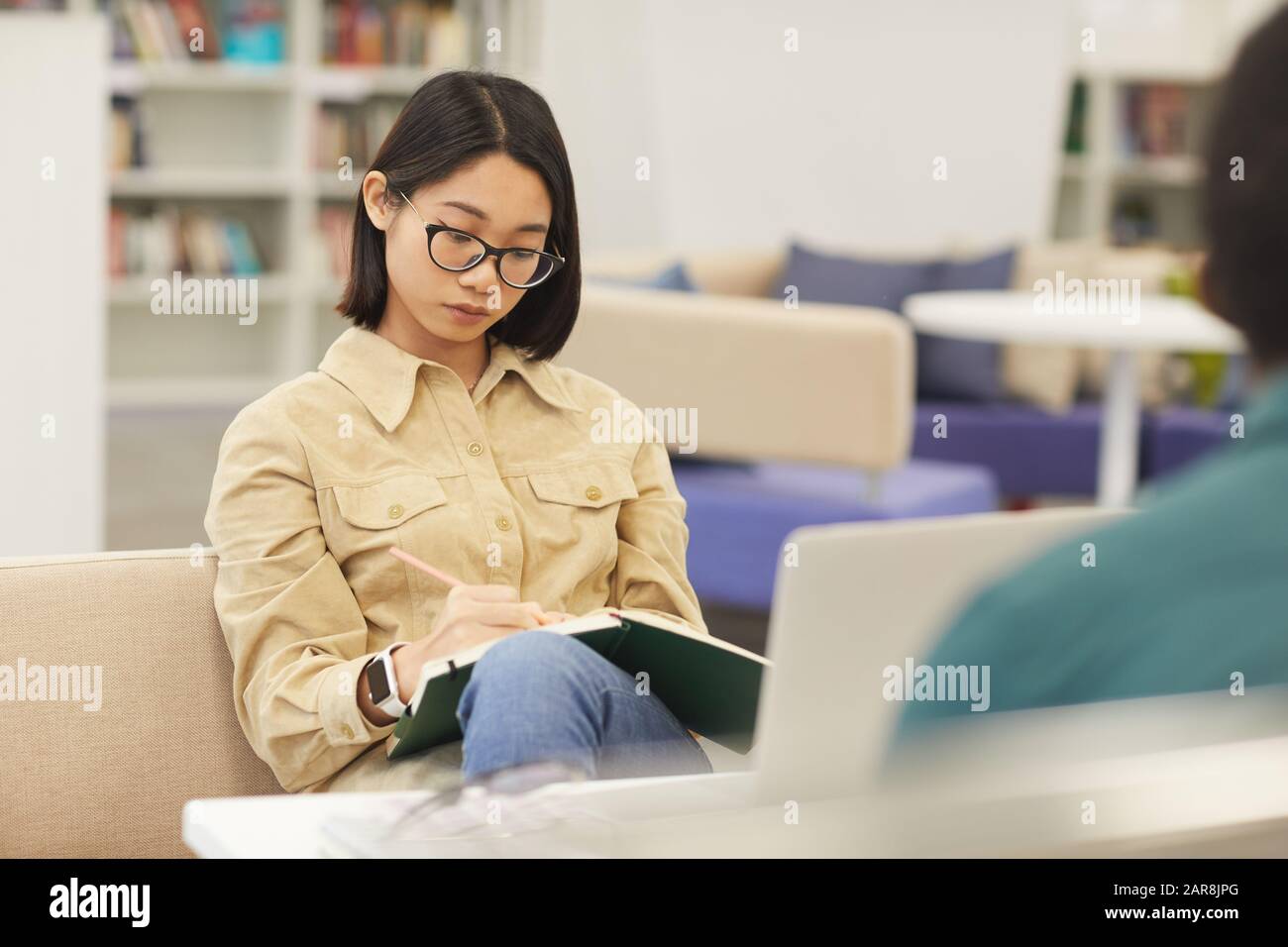 Portrait of Asian teenage girl studying in college library, writing in notebook while sitting on comfortable couch, copy space Stock Photo