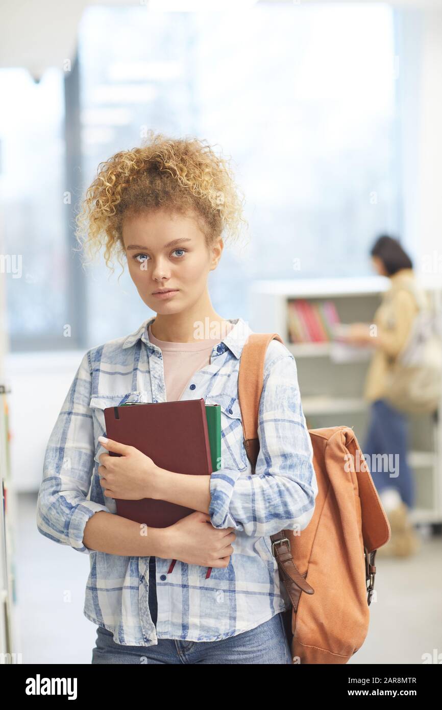 Waist up portrait of blonde female student holding book and looking at camera while posing by shelves in college library Stock Photo