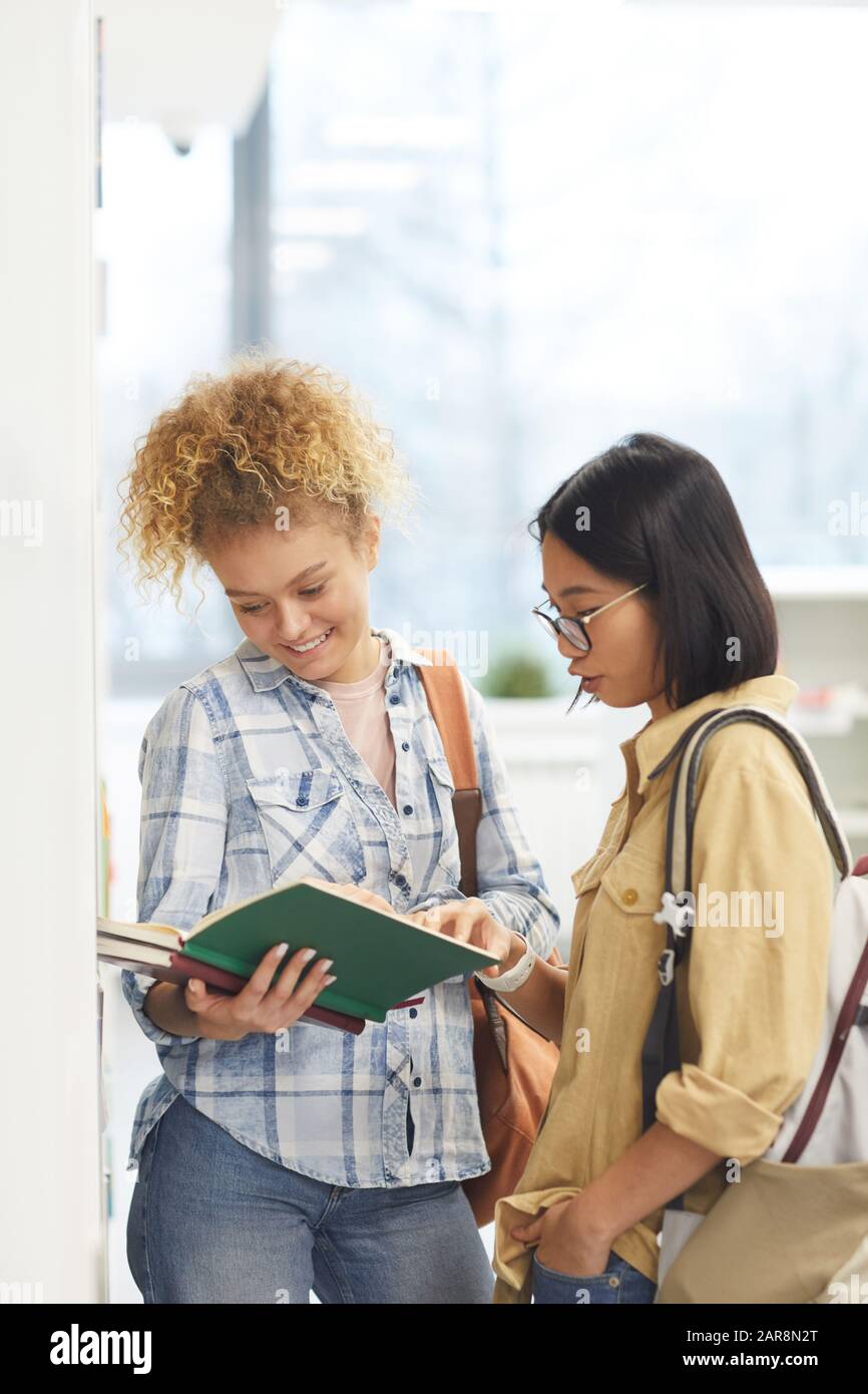 Waist up portrait of cheerful two female students reading book while standing by shelves in college library Stock Photo