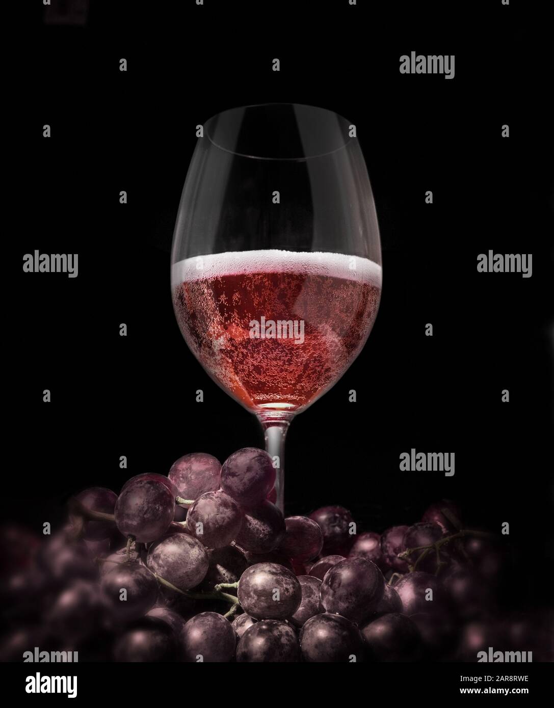 Surrounded by red grapes, a wine glass containing mad wine (sparkling and foam)or some grape beverage..low key, copy-space. Stock Photo