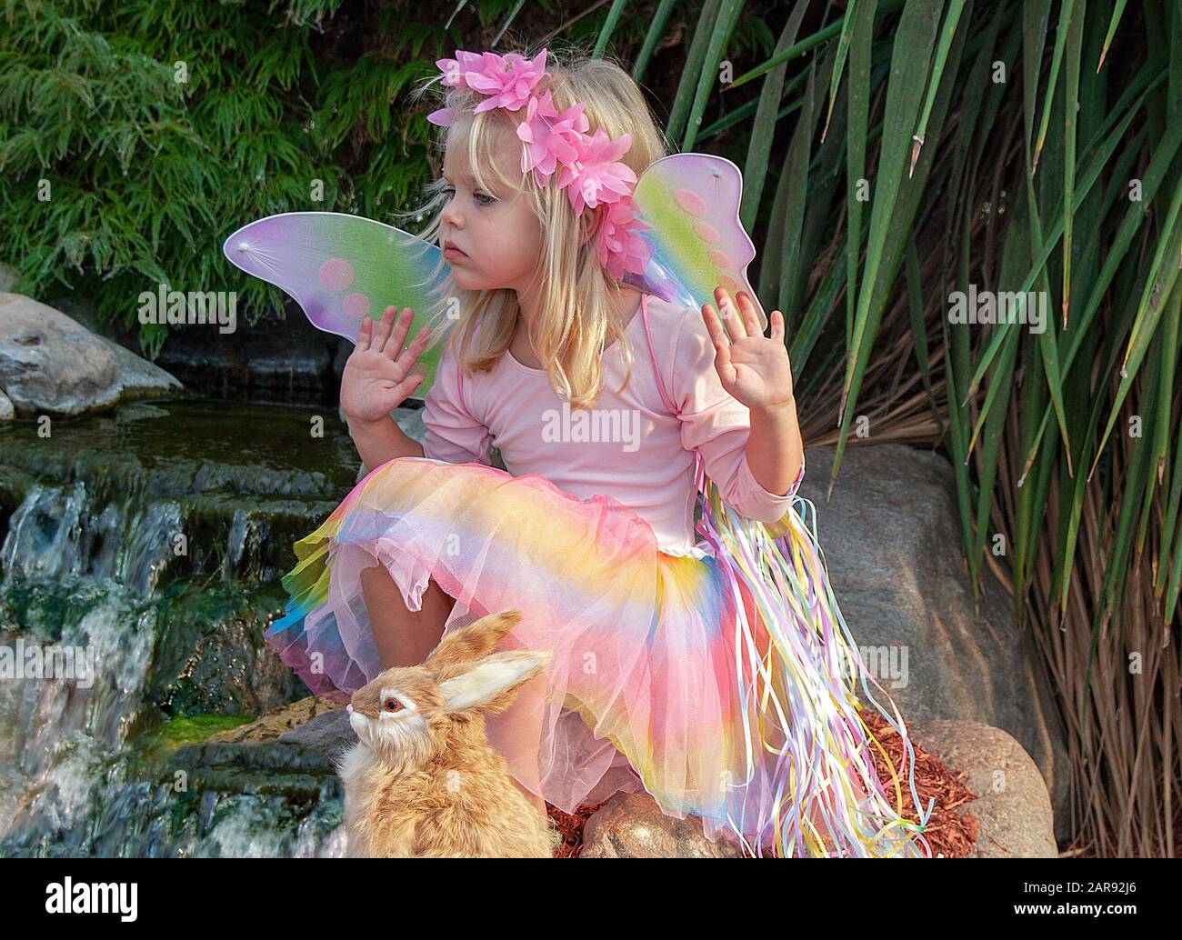 young blond Caucasian girl wearing a pastel rainbow fairy costume with wings sitting by waterfall in summer garden with rabbit Stock Photo
