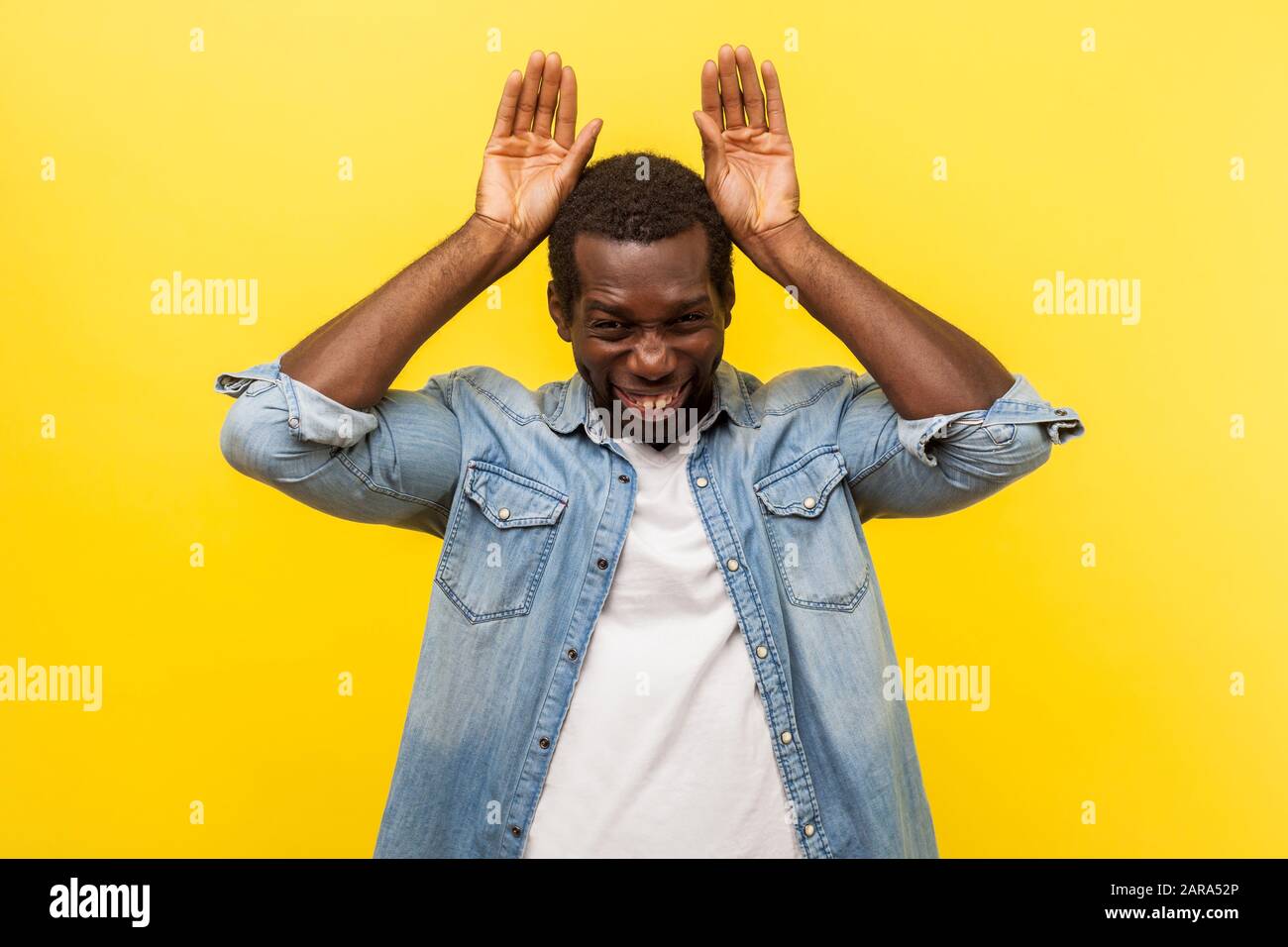 Portrait of extremely joyous man in denim casual shirt with rolled up sleeves showing bunny ears gesture, having fun with silly hilarious expression. Stock Photo
