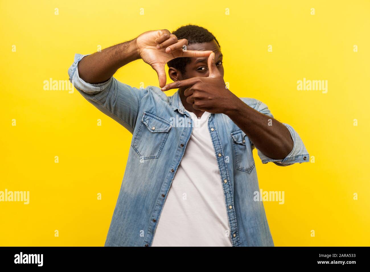 Portrait of focused curious man in denim casual shirt with rolled up sleeves looking through photo frame made of hands, viewing distant with interest. Stock Photo