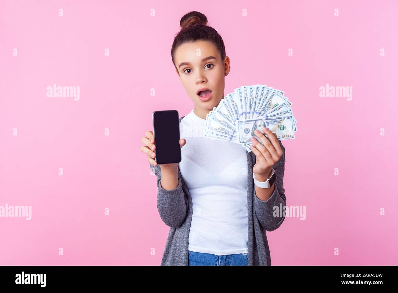 Online money transfer! Portrait of amazed brunette teen girl with bun hairstyle in casual clothes showing cell phone and dollars with surprised shocke Stock Photo