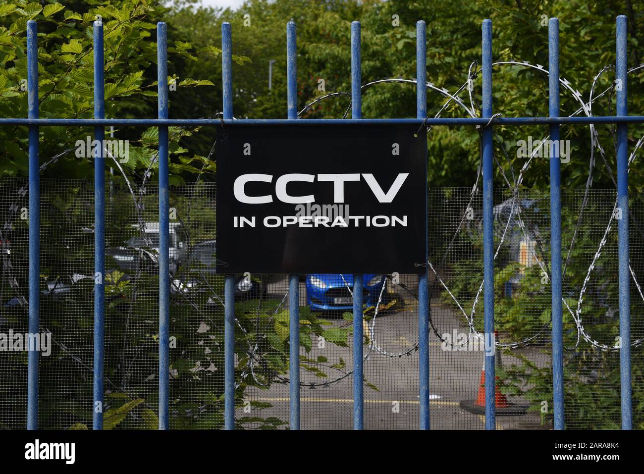 CCTV sign on a glue fence with razor wire. Protecting a car park with lots of trees. Stock Photo