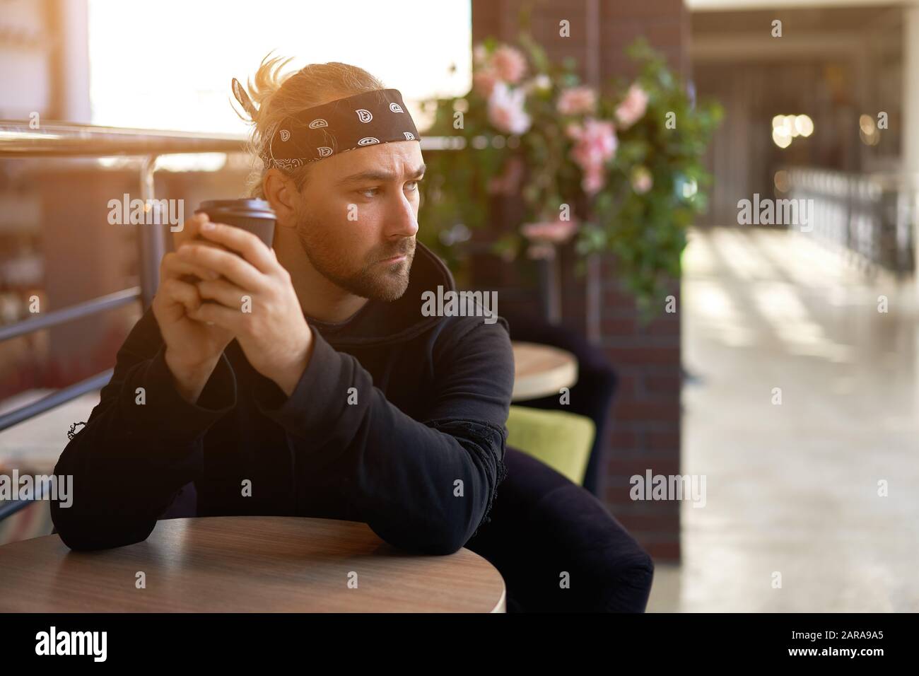Young handsome lonely man sits cafe table sad waiting for meeting drinking  coffee from a paper cup. Caucasian stylish guy dressed black hoodie bandana  Stock Photo - Alamy