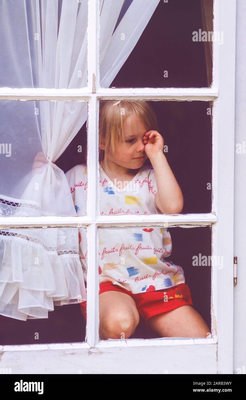 young girl stuck indoors looking out of a window rubbing her eyes