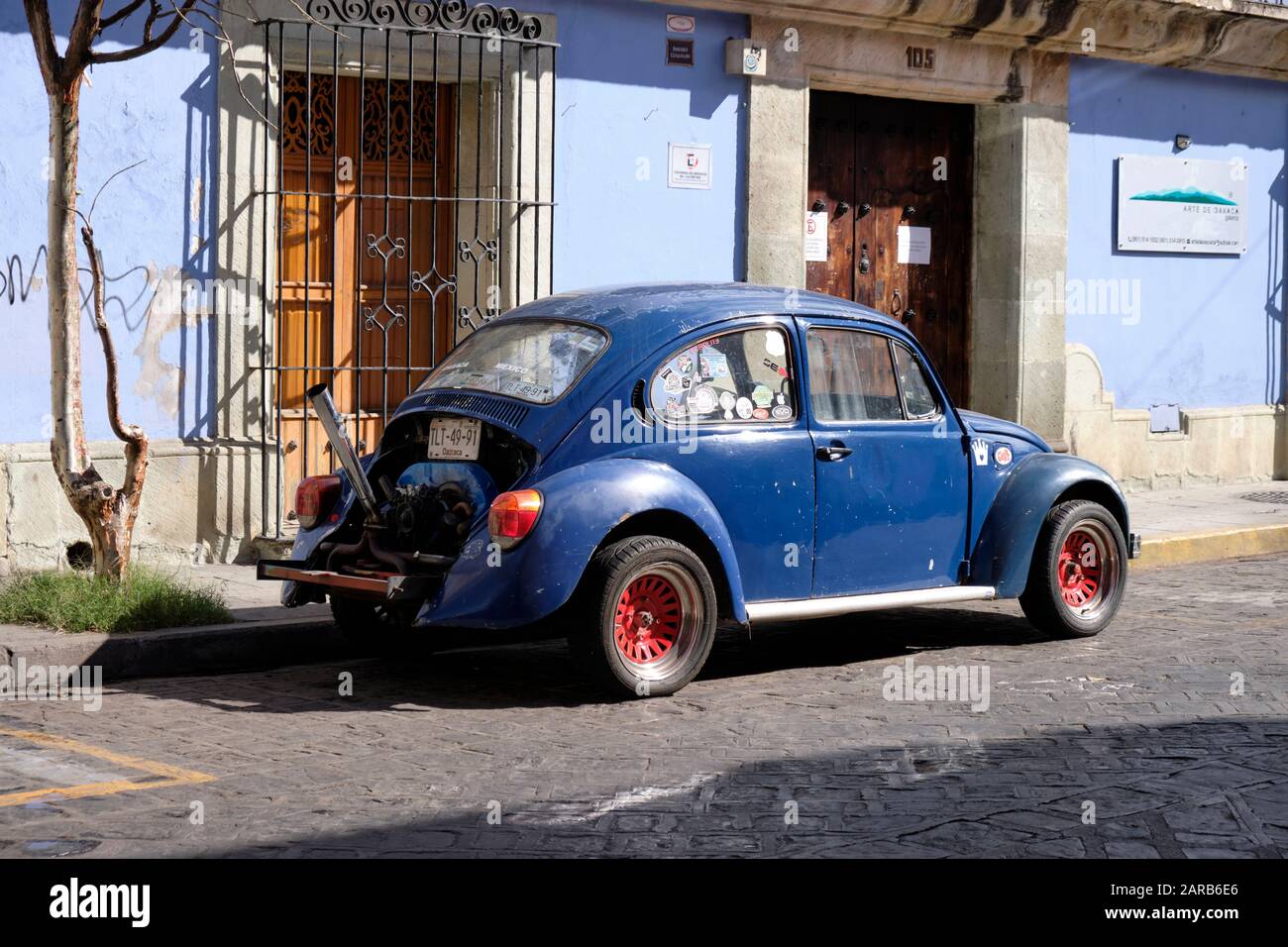 Blue classic Volkswagen beetle car parked on street next to a colonial house, painted also blue.  The engine has been modified with the hood removed Stock Photo