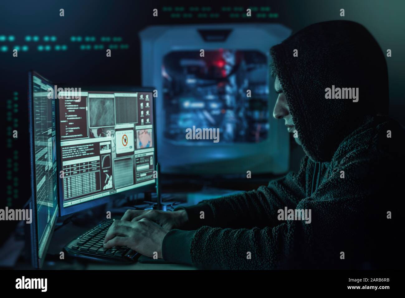 Dangerous hooded hacker in his hideout place which has a dark atmosphere, multiple displays Stock Photo