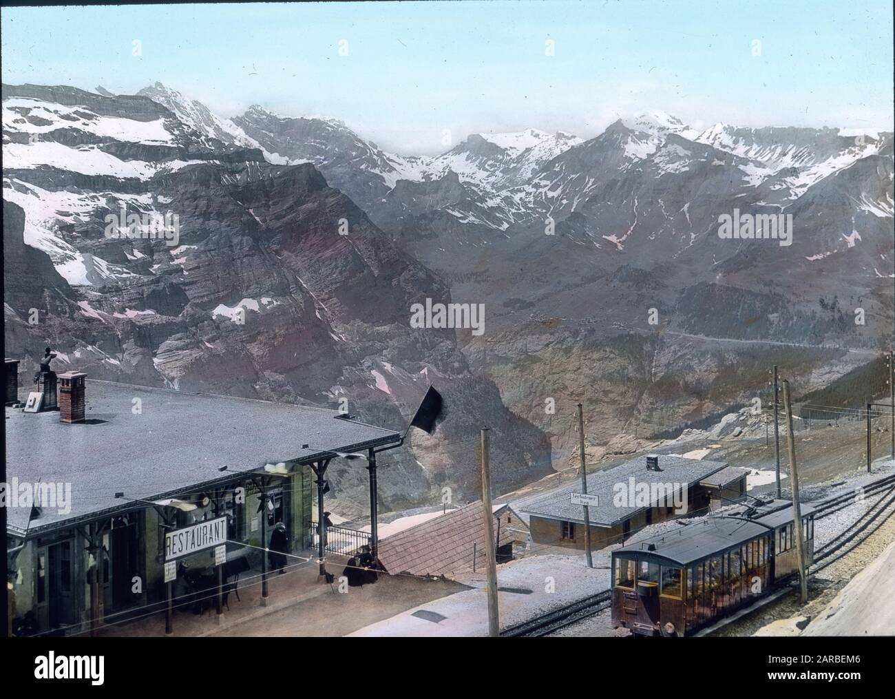 Die erste Haltestelle ist die Station Eigergletscher mit prachtvollem Blick auf die Berge des Lauterbrunnentales und Mürren. In wenigen Minuten läßt sich von hier aus die künstlicheEishöhle im Eigergletscher besuchen und dieser selbst betreten; in seinem unteren Ende ist er auch ohne Führer gefahrlos zu begehen. , history, historical, Switzerland, travel, 1910s, 20th century, Carl Simon, mountain, virgin, Jungfraubahn, Station Eigergletscher, glacier, Bernese upland, Carl Simon, hand coloured glass slide Stock Photo