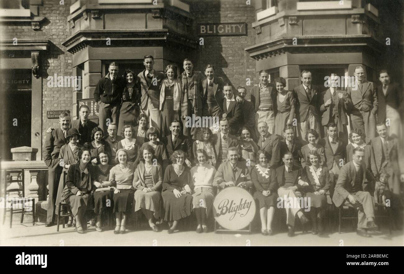 A group posing outside a guest house going by the name of 'Blighty' around what looks to be a dinner gong emblazoned with the name.     Date: 1930s Stock Photo