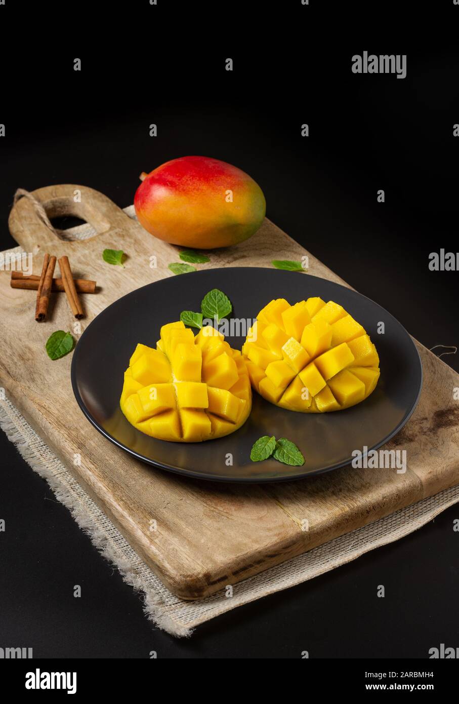 Mango cut in half and cubed on a wooden board, a whole mango and cinnamon branches, black background with space for copying Stock Photo