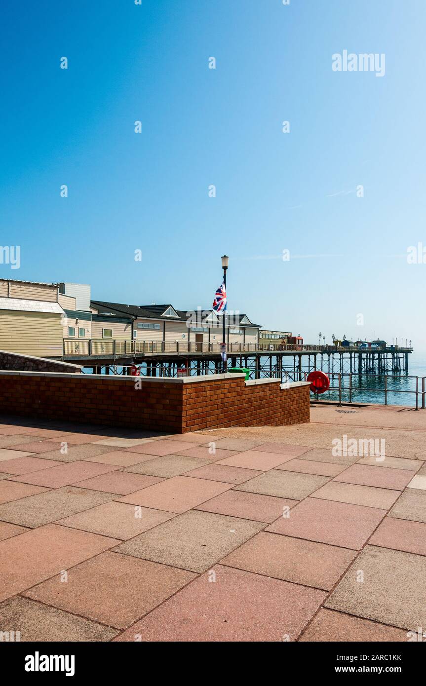 An area paved with coloured concrete slabs and bordered by a red brick wall leads down a wide ramp to the promenade close by Teignmouth Grand Pier Stock Photo