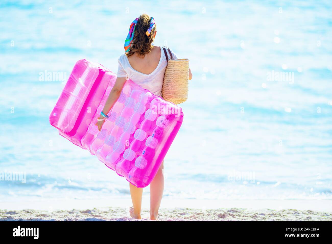 Seen from behind trendy woman in white t-shirt and pink shorts with beach straw bag on the beach holding inflatable mattress. Stock Photo