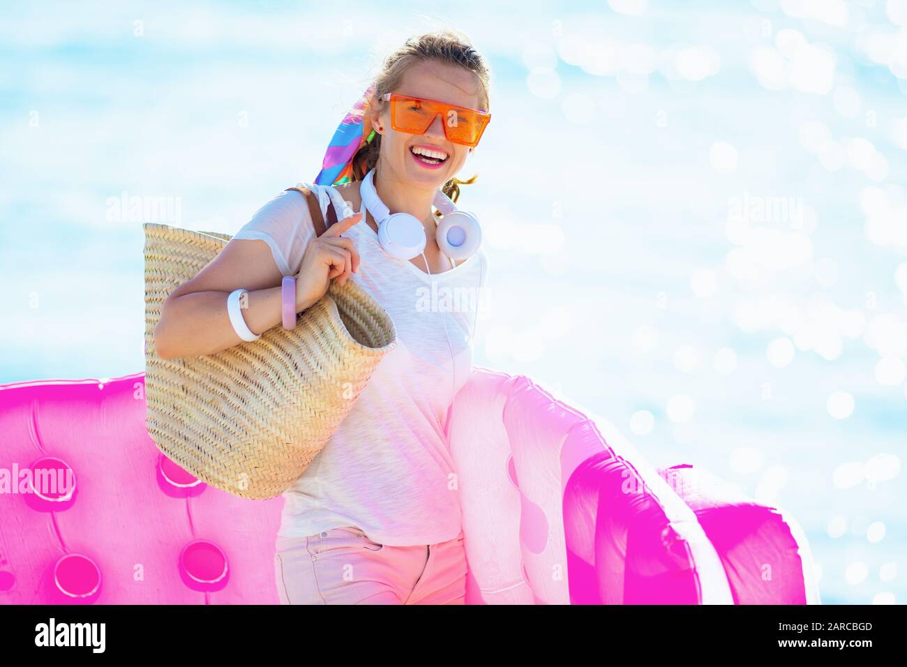 Portrait of smiling trendy woman in white t-shirt and pink shorts with beach straw bag on the seacoast holding inflatable mattress. Stock Photo