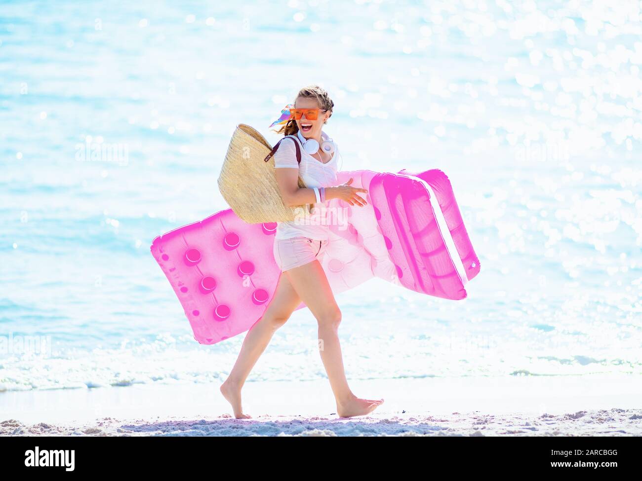 happy modern woman in white t-shirt and pink shorts with beach straw bag on the seashore holding inflatable mattress and walking. Stock Photo