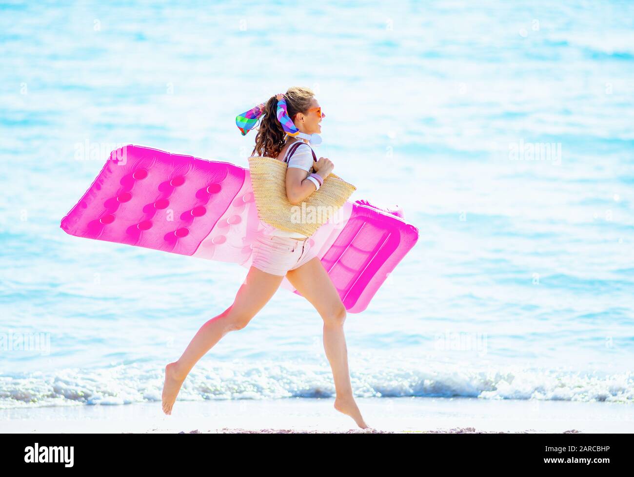 happy young woman in white t-shirt and pink shorts with beach straw bag on the seacoast holding inflatable mattress and running. Stock Photo