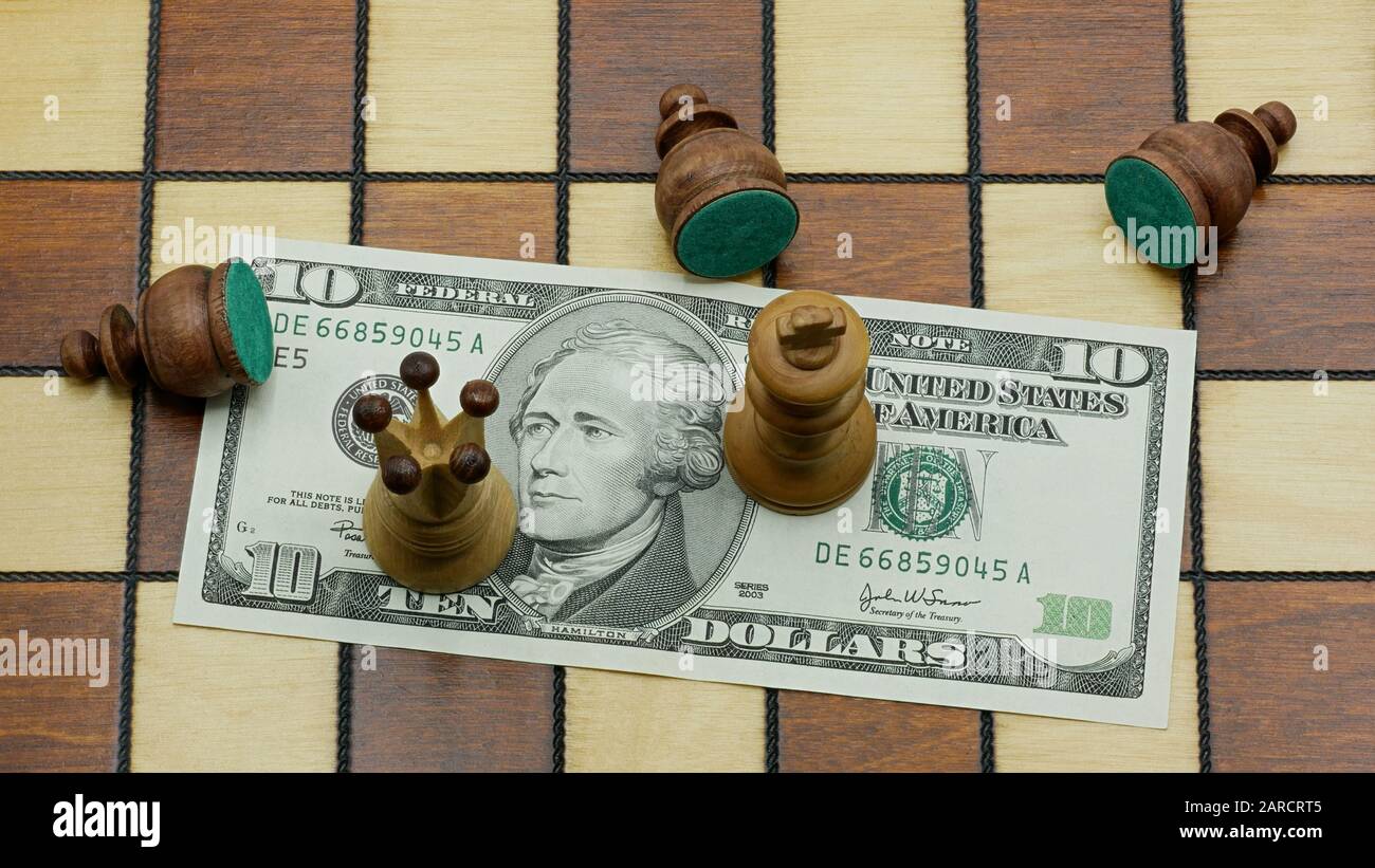 Money Rules: Defeated Black Pawn Chess Pieces Lying Around A White King And A White Queen Standing On A 10 Dollars Banknote On A Chess Board Stock Photo