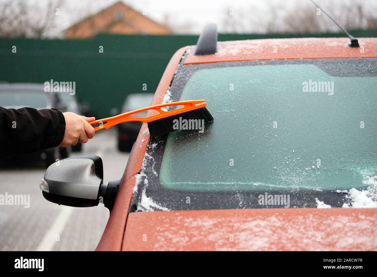 Man clears snow from icy windows of car. Brush in mans hand. Windshield of orange auto, horizontal view. Stock Photo