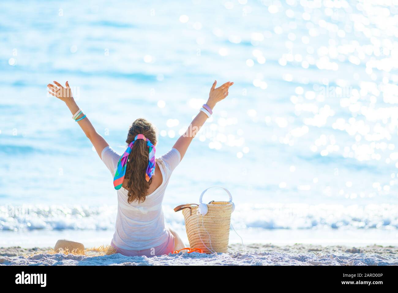 Seen from behind modern woman in white t-shirt and pink shorts with headphones and beach straw bag sitting and rejoicing on the ocean coast. Stock Photo