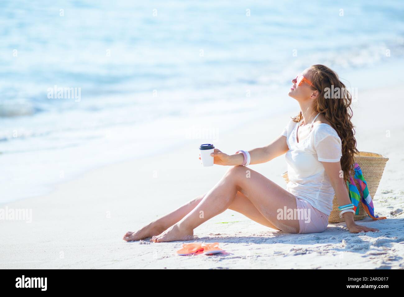 relaxed modern woman in white t-shirt and pink shorts with coffee cup sitting on the seashore. Stock Photo