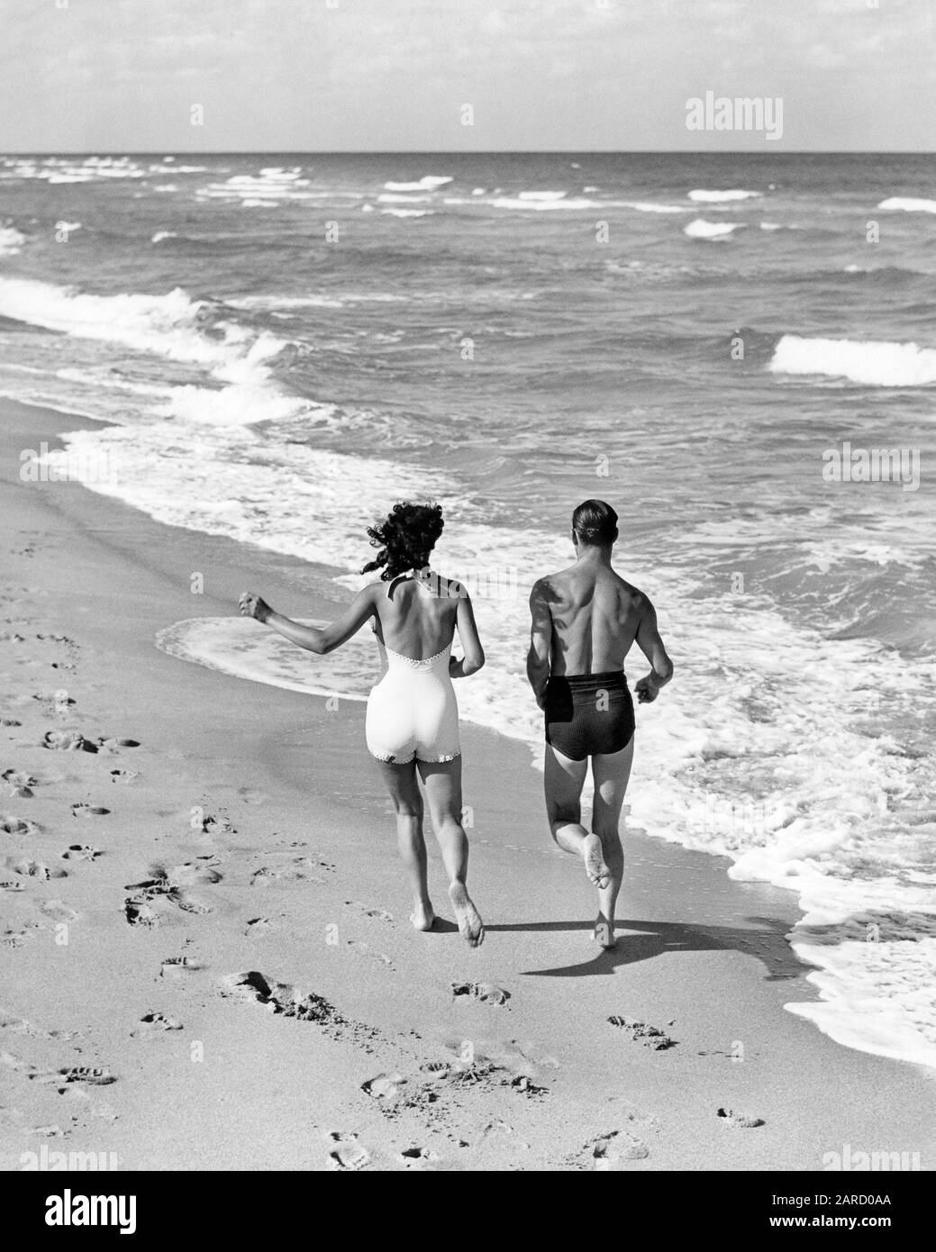 1930s 1940s COUPLE WEARING BATHING SUITS JOGGING RUNNING TOGETHER AT EDGE OF SURF ON OCEAN BEACH - b7071 HAR001 HARS MID-ADULT MAN MID-ADULT WOMAN BLACK AND WHITE CAUCASIAN ETHNICITY HAR001 OLD FASHIONED Stock Photo