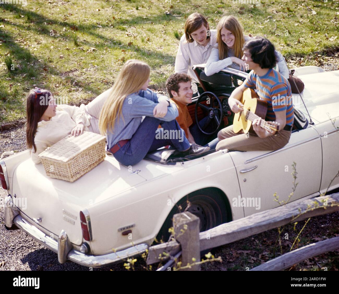 1960s GROUP 6 STYLISH TEENS ON WHITE TRIUMPH CONVERTIBLE WITH PICNIC 1960s-group-6-stylish-teens-on-white-triumph-convertible-with-picnic