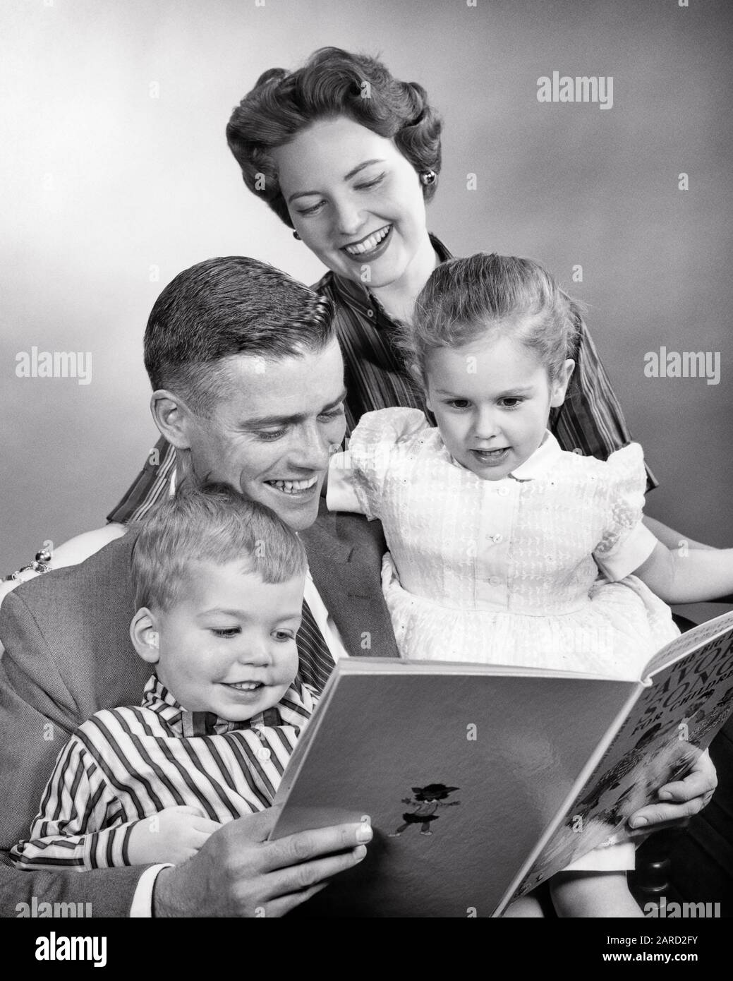 1950s FAMILY AT HOME WOMAN MAN GIRL BOY SMILING MOTHER WATCHING LOVING FATHER READ BOOK TO DAUGHTER AND SON - r506 HAR001 HARS INDOORS NOSTALGIC PAIR 4 SUBURBAN URBAN MOTHERS OLD TIME BUSY FUTURE NOSTALGIA BROTHER READ OLD FASHION SISTER 1 JUVENILE COMMUNICATION SECURITY TEAMWORK INFORMATION SONS PLEASED FAMILIES JOY LIFESTYLE FEMALES MARRIED BROTHERS SPOUSE HUSBANDS HEALTHINESS HOME LIFE COPY SPACE HALF-LENGTH LADIES DAUGHTERS PERSONS CARING MALES SERENITY SIBLINGS CONFIDENCE SISTERS FATHERS B&W PARTNER SUCCESS HAPPINESS WELLNESS HEAD AND SHOULDERS CHEERFUL DISCOVERY LEISURE STRENGTH AND DADS Stock Photo