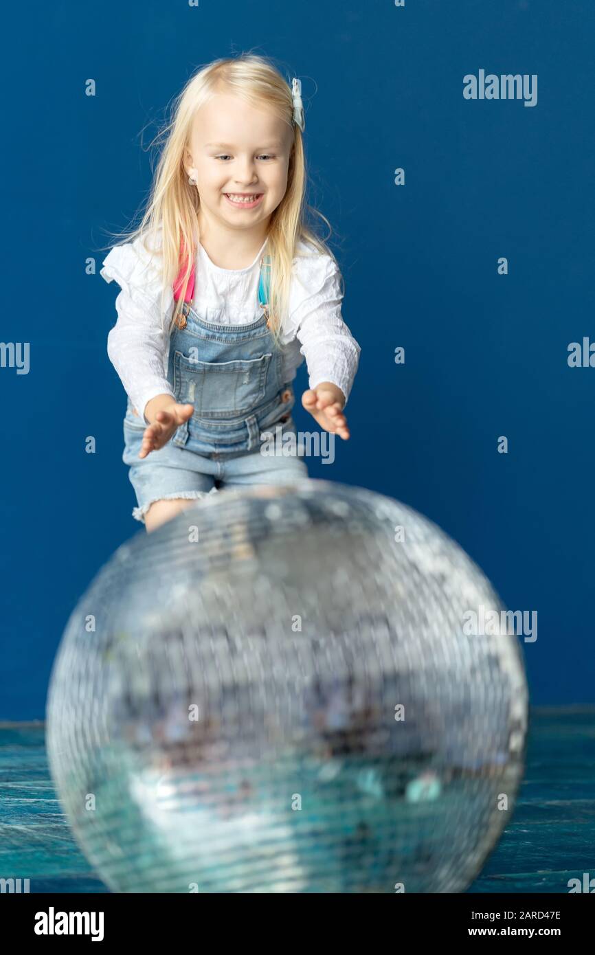 Happy toddler blonde girl havig fun while rolling giant disco ball on the floor Stock Photo