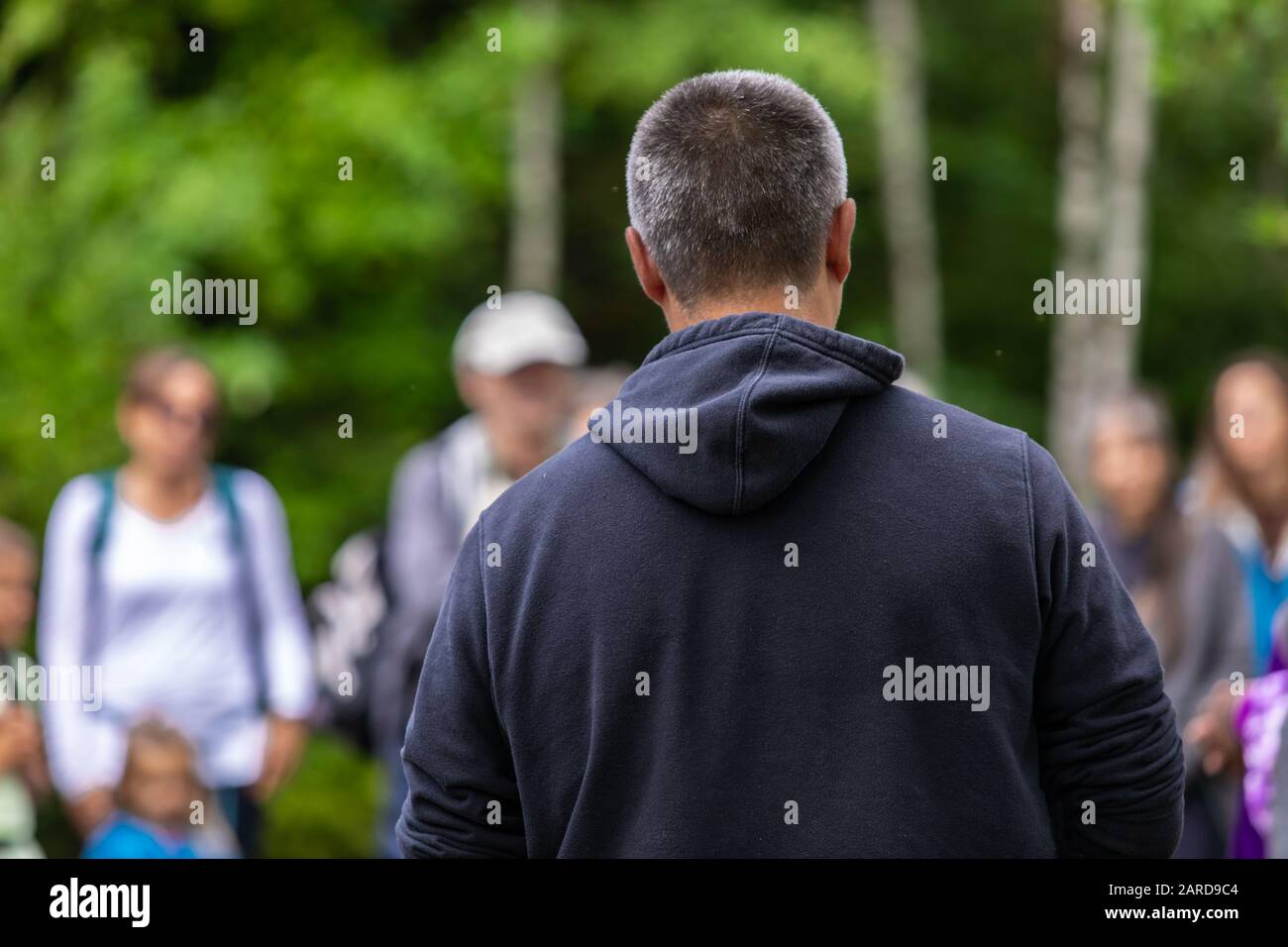 Rear view of male wearing blue warm clothing hoodie and telling fairytale story to people against trees in festival during event Stock Photo