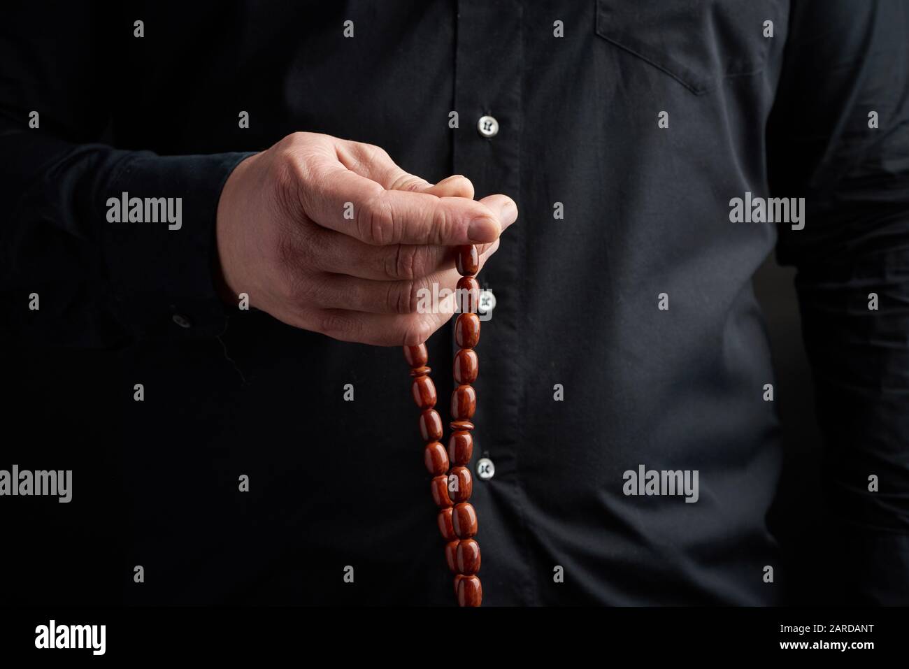 man in a black shirt holds a brown stone rosary in his left hand, low key. used to count prayers or other ritual actions, to preserve attention and co Stock Photo