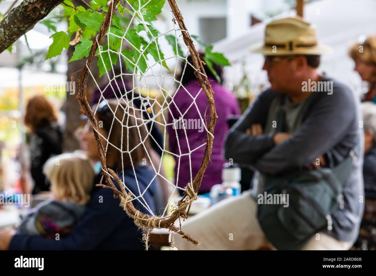 Closeup of handmade dreamcatcher hanging on tree branch with defocus group of people sitting and looking at something at outdoor event Stock Photo