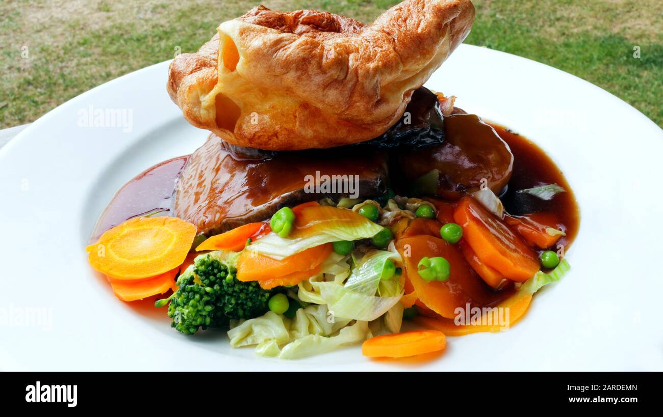 Traditional English roast with Yorkshire pudding served with sliced beef, carrots, peas, broccoli and gravy, on a white plate . Stock Photo