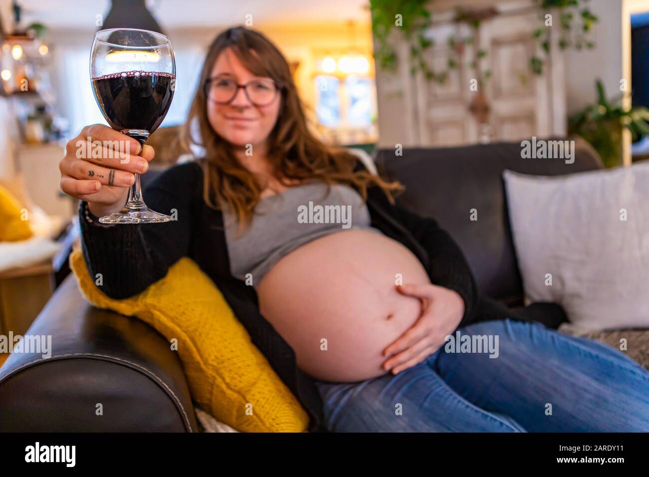 A shallow depth of field as a blurry pregnant woman relaxes on sofa in family room with glass of red wine, risking fetal alcohol syndrome. Copy space to right Stock Photo