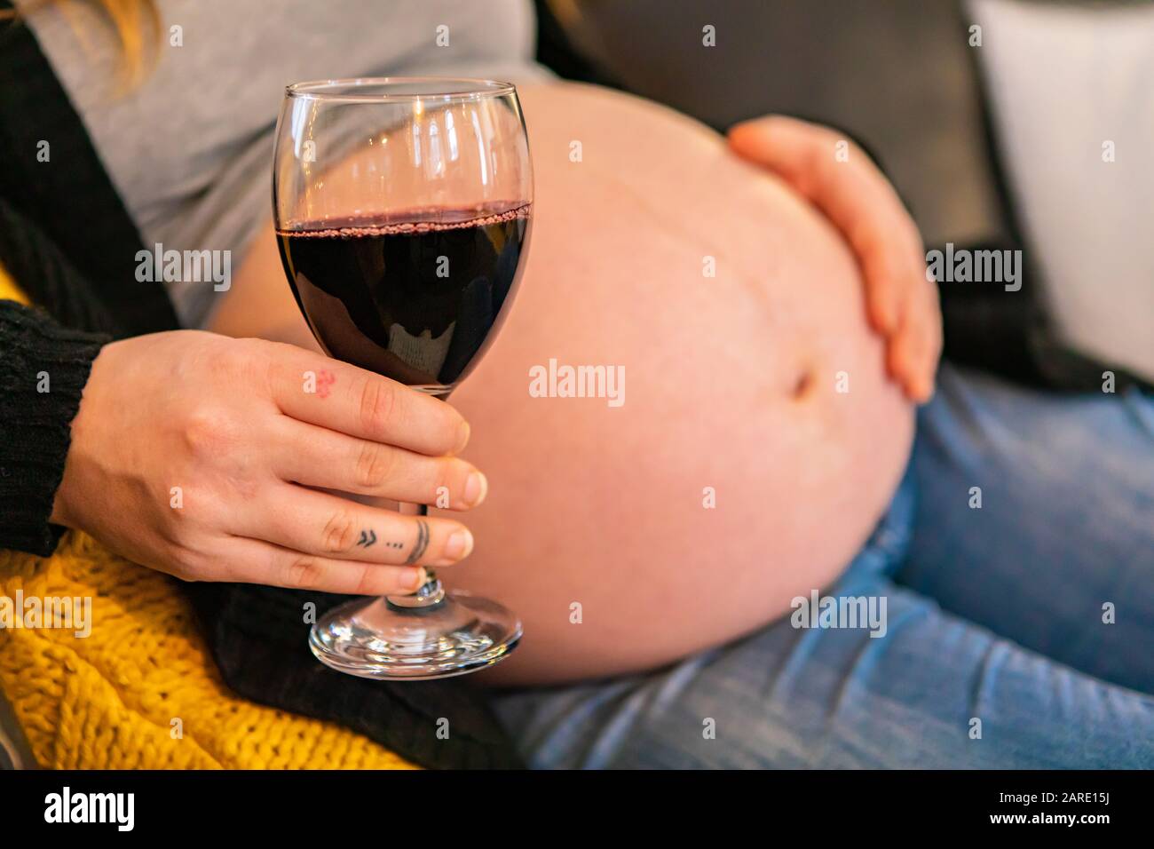 Closeup shot of a glass of red wine in hand of a woman in later stages of pregnancy, with swollen baby bump blurry in background, risk to unborn child Stock Photo